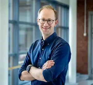 Leende man med armarna i kors, glasögon och blå skjorta står framför ett fönster.