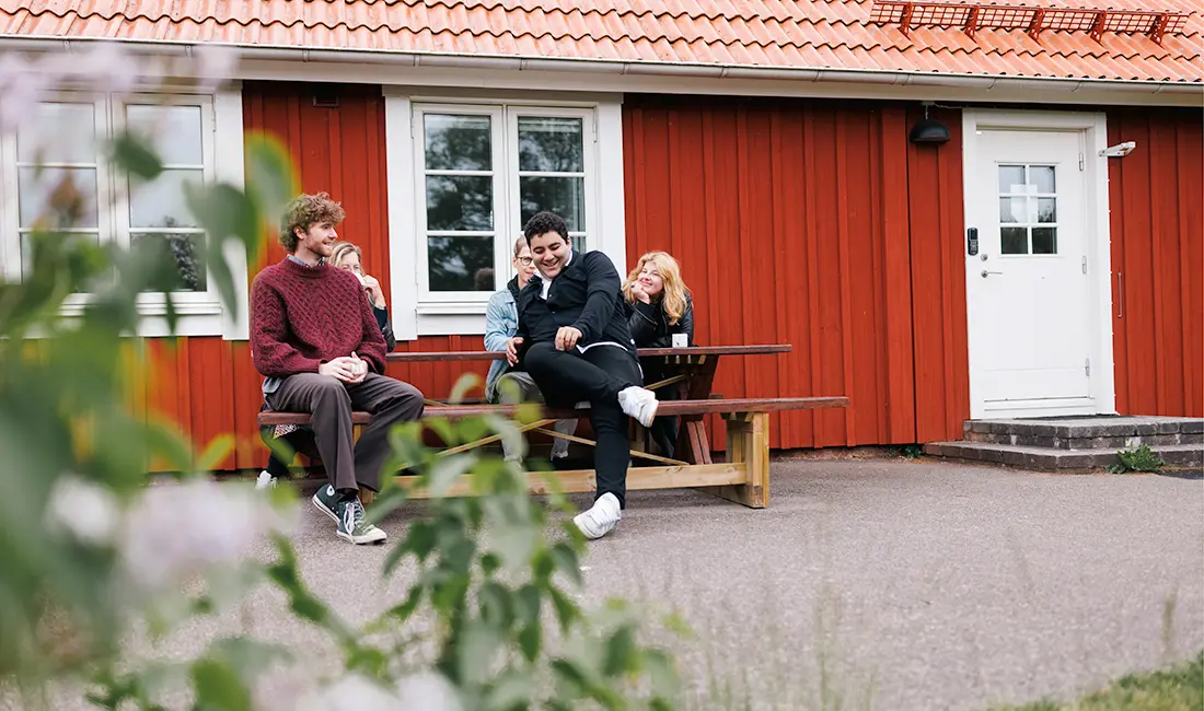 Fem personer sitter på en träbänk utanför ett rött trähus.