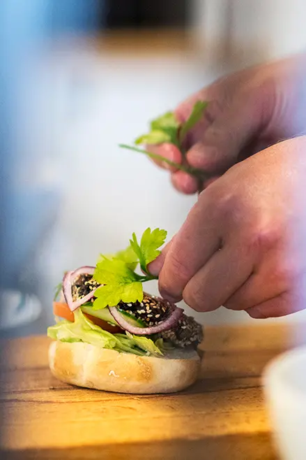 Närbild av händer som placerar sallad på en hamburgare.