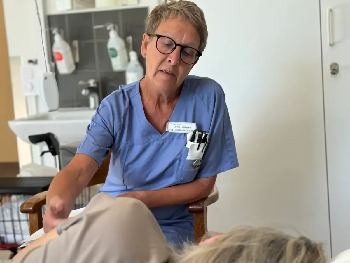 Sjuksköterksa i blå skjorta sitter vid sidan av en säng. Klappar på patient.