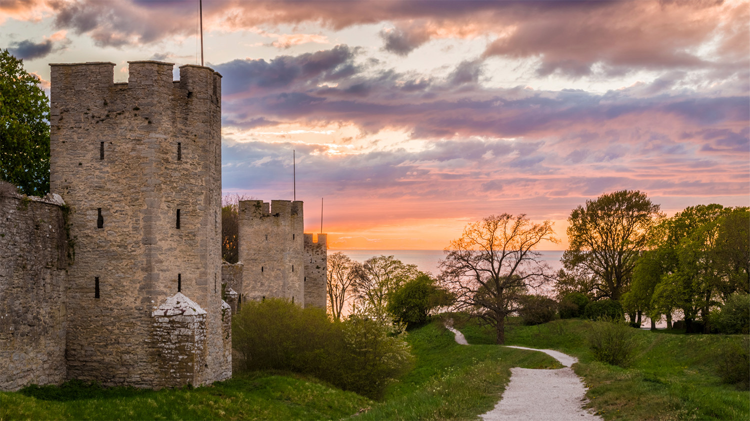 Visby ringmur och en stig ner mot havet, belyst av kvällsljus som färgar molnen i pastellfärger.