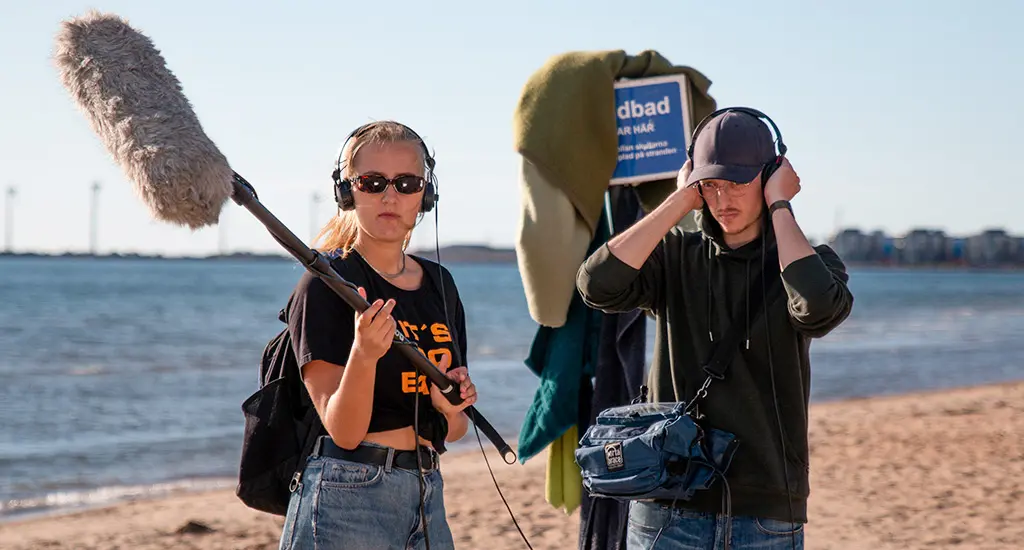 Två unga personer står på en strand och håller i ljudutrustning på en inspelning.