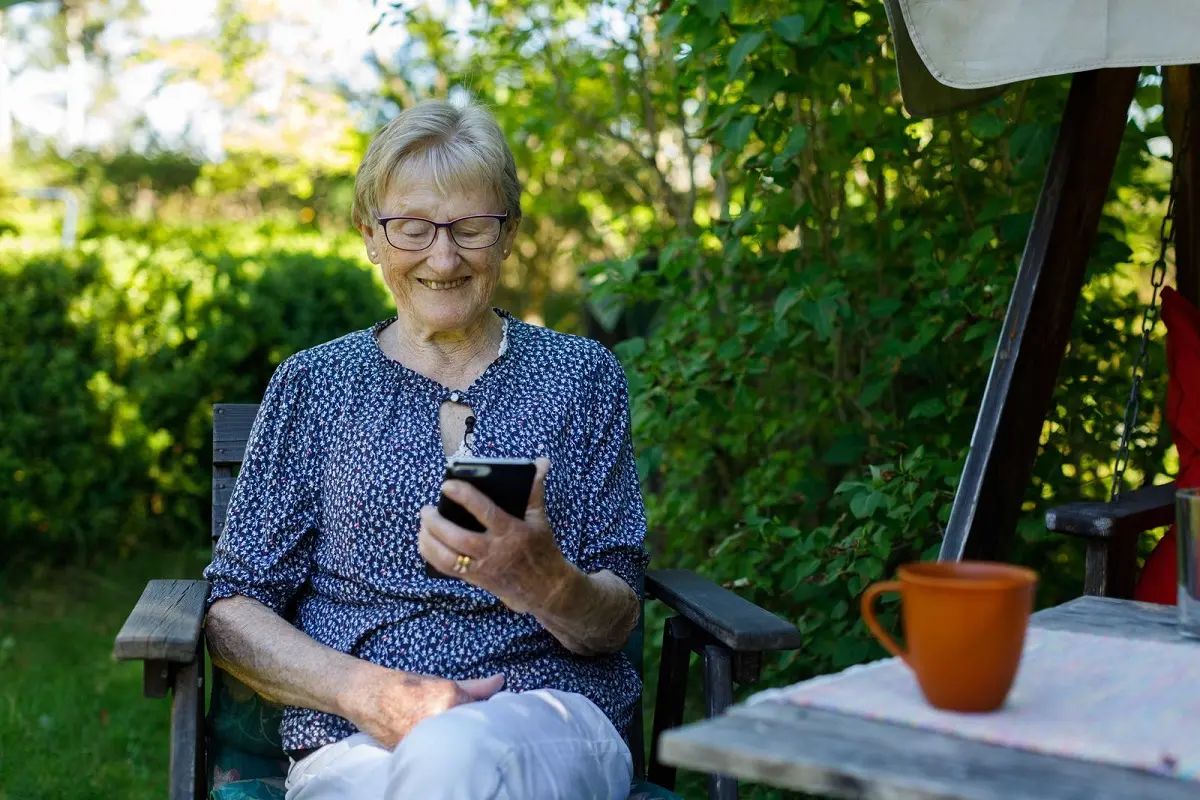 Äldre person sitter utomhus vid ett bord och tittar på en mobiltelefon som hon håller i handen.