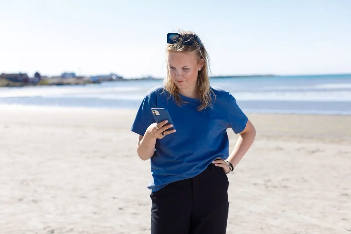 Ung person står på en strand, tittar på mobiltelefon som hon håller i handen.