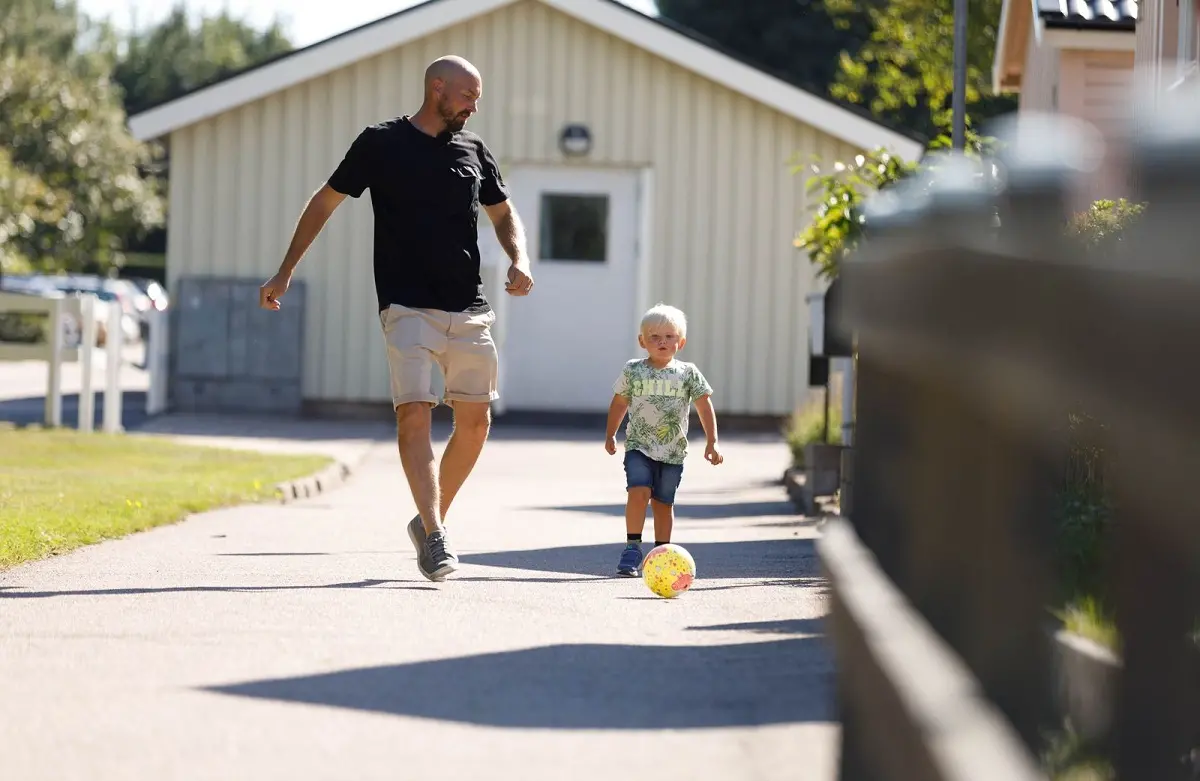 Vuxen person spelar fotboll med ett barn på en gata i ett bostadsområde.