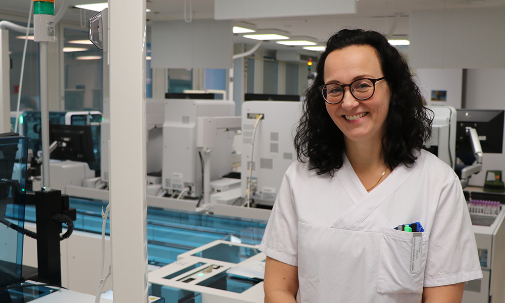 Leende kvinna med vit rock, mörkt hår och glasögon står i en laboratoriemiljö