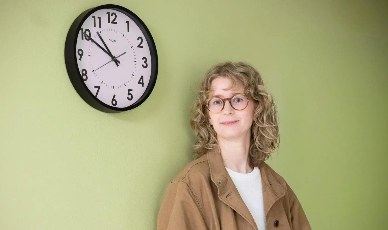 En blond kvinna står framför en grön vägg med en klocka. 