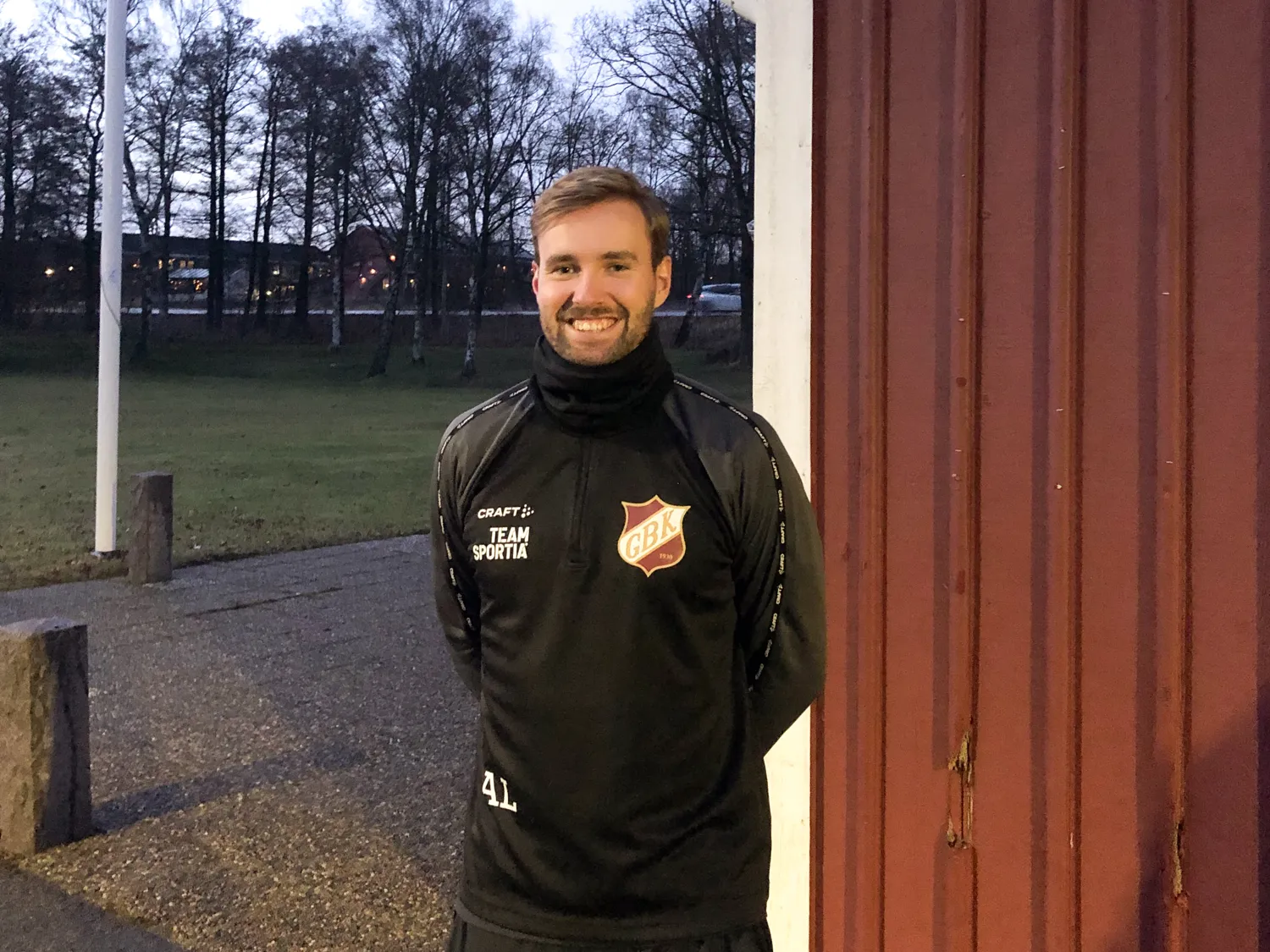 En kille i träningsoverall står vid en röd byggnad