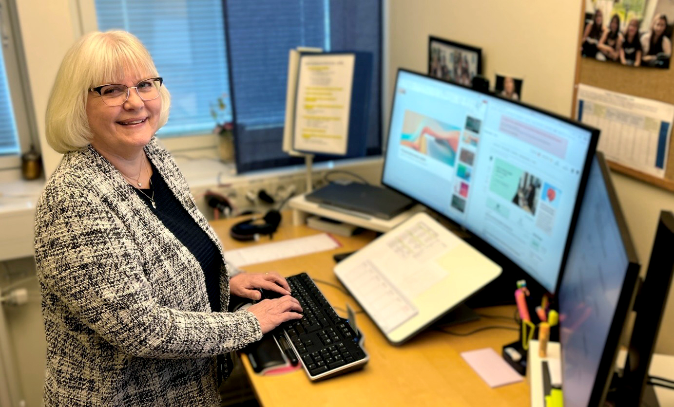 Anki står vid ett höj- och sänkbart skrivbord på ett kontor och arbetar vid flera datorskärmar. Hon tittar i kameran och ler.
