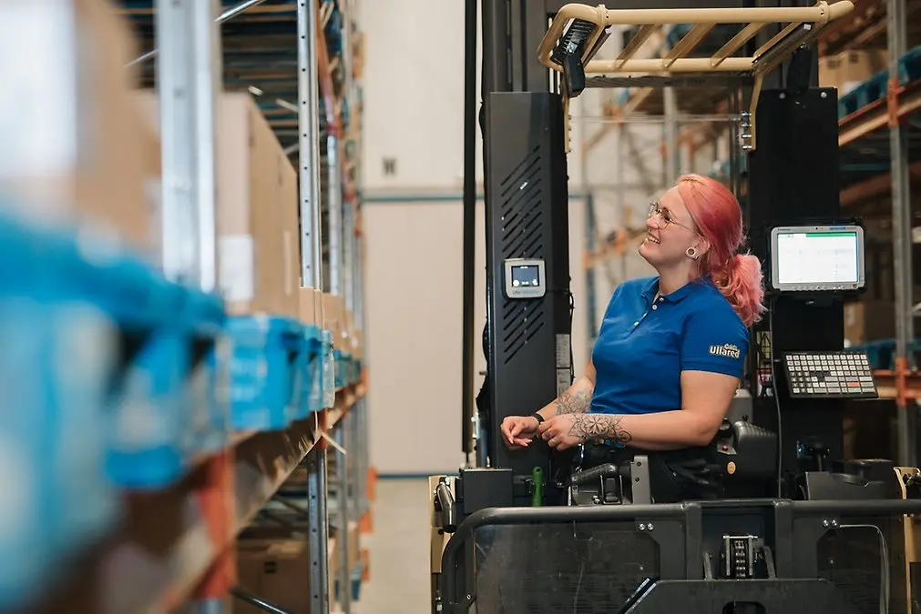 Kvinna med rosa hår och blå arbetsskjorta kör truck i lagermiljö