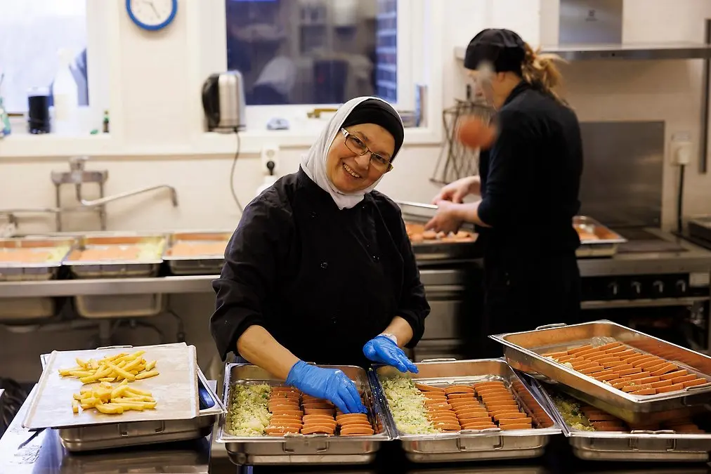 Leende kvinna i svarta kläder, vit sjal, glasögon och blå handskar lägger upp mat i tråg.