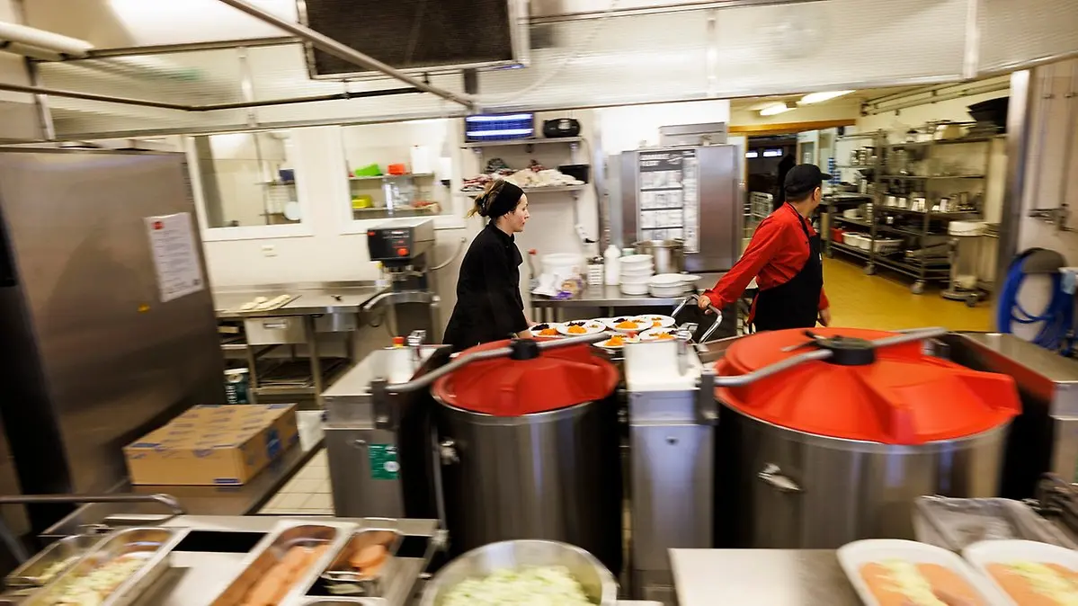 Inne i skolköket, stora kastruller i förgrunden, två personer som 