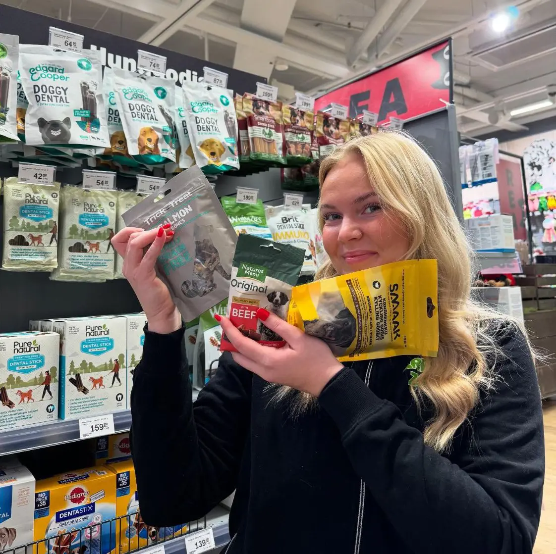 En ung person håller upp hundgodispåsar i en djuraffär. Hyllor med produkter i bakgrunden.