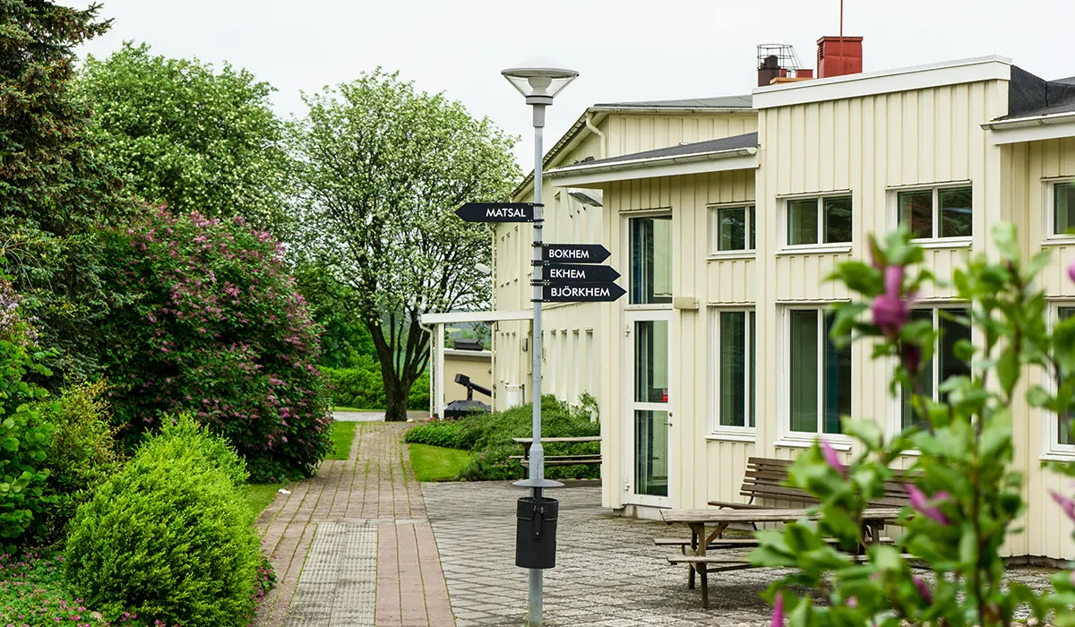 Utsidan av ett gult enplanshus. Framför huset finns träd och buskar.