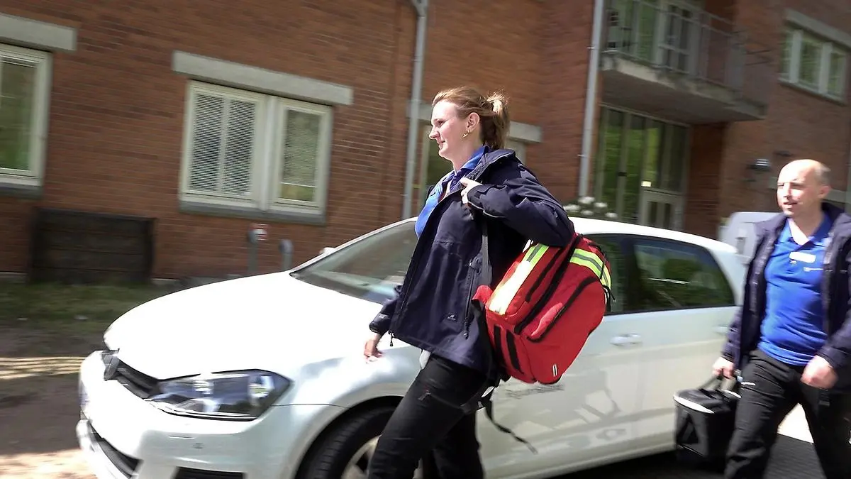 Två personer i vårdkläder går på en gata, bär på var sin väska.