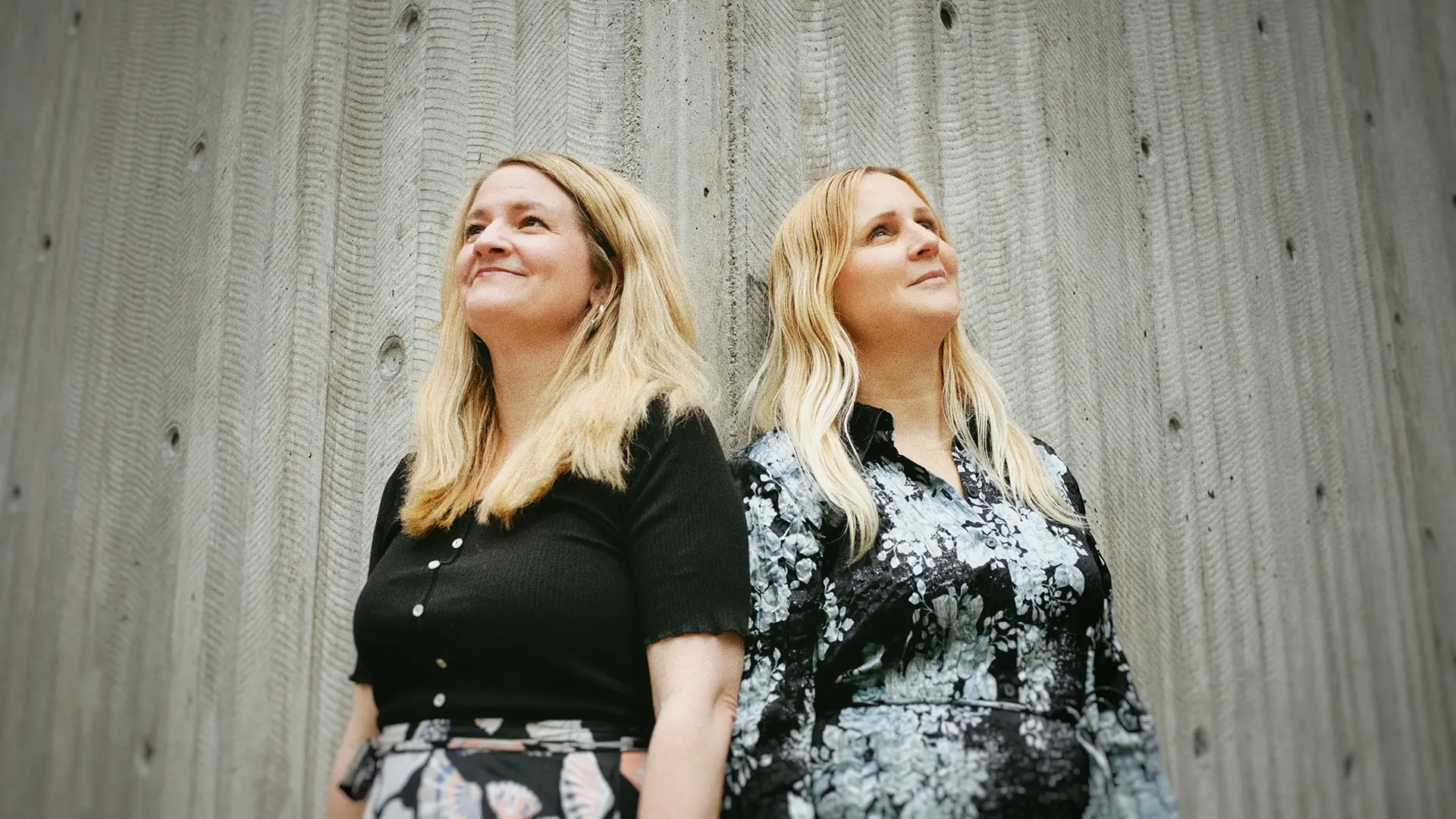 Två kvinnor med långt ljust hår, står bredvid varandra och tittar utåt och uppåt.