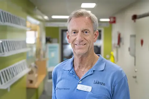 Man med grått hår och blå sjukhuskläder, står i en korridor