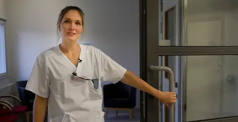 Kvinna i vita vårdkläder håller upp en dörr.
