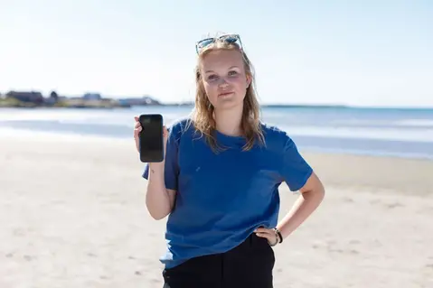 Ung person står på en strand och håller upp en mobiltelefon.