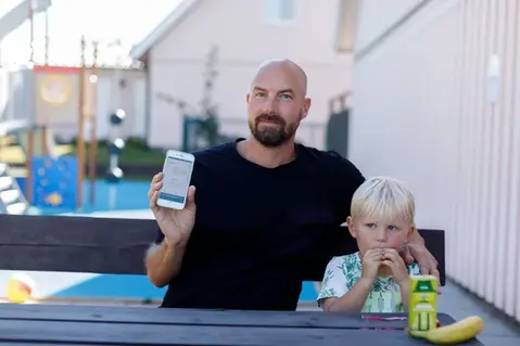 Vuxen person sitter på en bänk med ett litet barn bredvid sig, håller upp en mobiltelefon.
