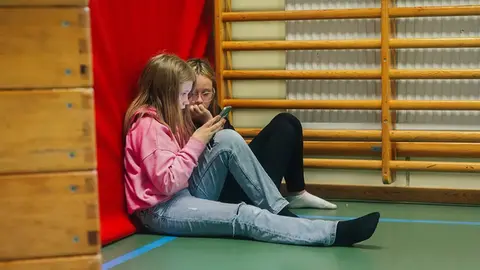 Två tjejer sitter på golvet i en gymnastiksal och tittar på en mobilskärm. Ribbstolar på väggen.