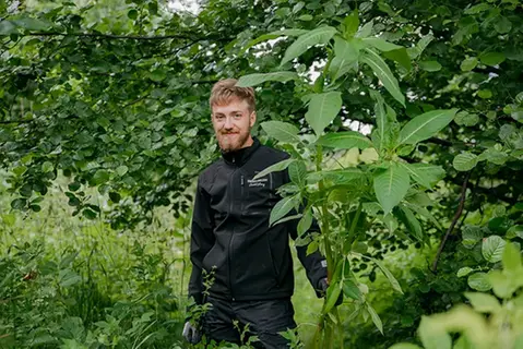 Kille i svarta kläder, mustasch och lite skägg står bland grönska