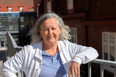 Kvinna med grått halvlångt hår och sjukhuskläder står utanför sjukhusbyggnad