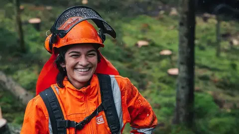 Ung, leende tjej i orangea skogsarbetarkläder och uppfällt visir står i en grön skog