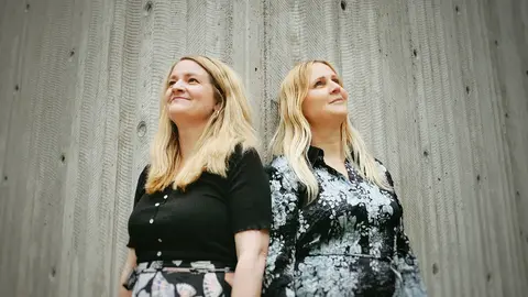 Två kvinnor med långt ljust hår, står bredvid varandra och tittar utåt och uppåt.